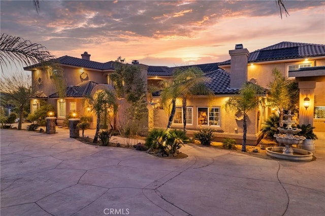 mediterranean / spanish-style home featuring a tile roof, roof mounted solar panels, stucco siding, a chimney, and driveway