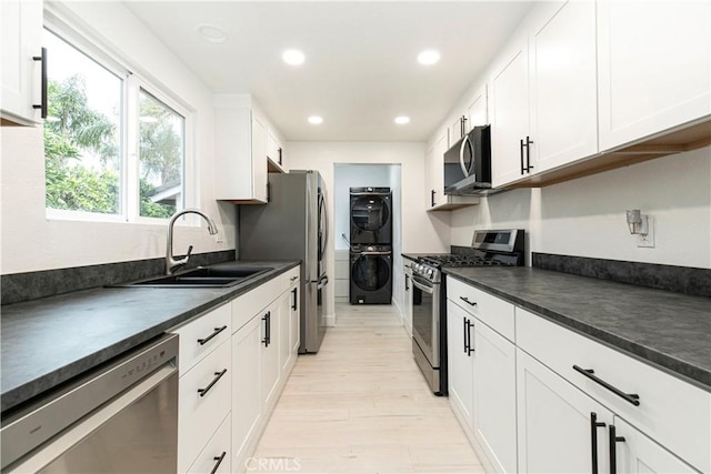 kitchen with white cabinets, dark countertops, appliances with stainless steel finishes, stacked washing maching and dryer, and a sink