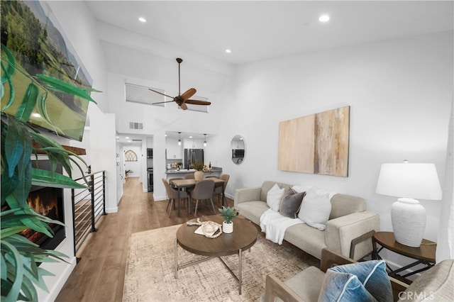 living room featuring a warm lit fireplace, visible vents, light wood-style flooring, ceiling fan, and high vaulted ceiling