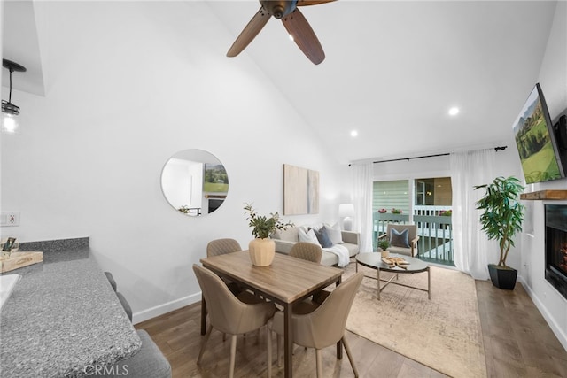 dining room featuring recessed lighting, wood finished floors, high vaulted ceiling, a warm lit fireplace, and baseboards