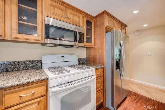 kitchen with appliances with stainless steel finishes, glass insert cabinets, light stone counters, and brown cabinets