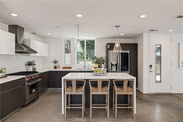 kitchen featuring a breakfast bar, stainless steel appliances, light countertops, wall chimney range hood, and concrete floors