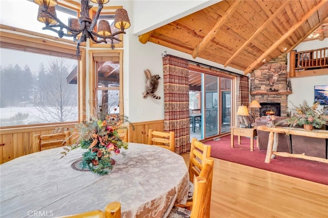 dining area featuring a wainscoted wall, vaulted ceiling with beams, an inviting chandelier, wood ceiling, and a stone fireplace