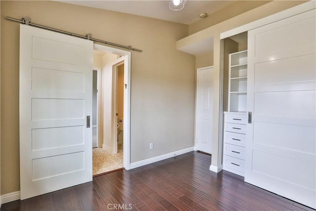 unfurnished bedroom featuring a barn door, baseboards, a closet, and dark wood finished floors