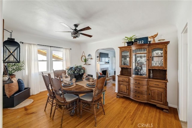 dining room featuring baseboards, arched walkways, a ceiling fan, light wood-type flooring, and a fireplace