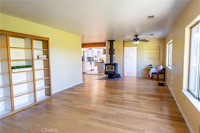 unfurnished living room featuring visible vents, a ceiling fan, a wood stove, light wood-type flooring, and baseboards