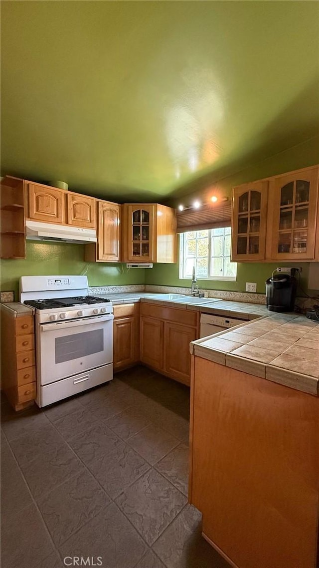 kitchen featuring white gas range, tile counters, glass insert cabinets, a sink, and under cabinet range hood