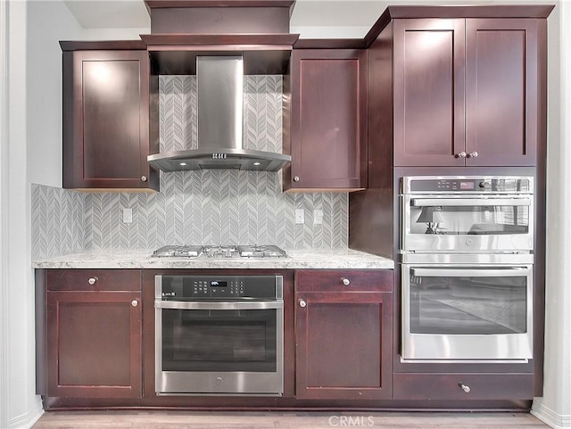 kitchen with stainless steel appliances, reddish brown cabinets, wall chimney range hood, decorative backsplash, and light stone countertops