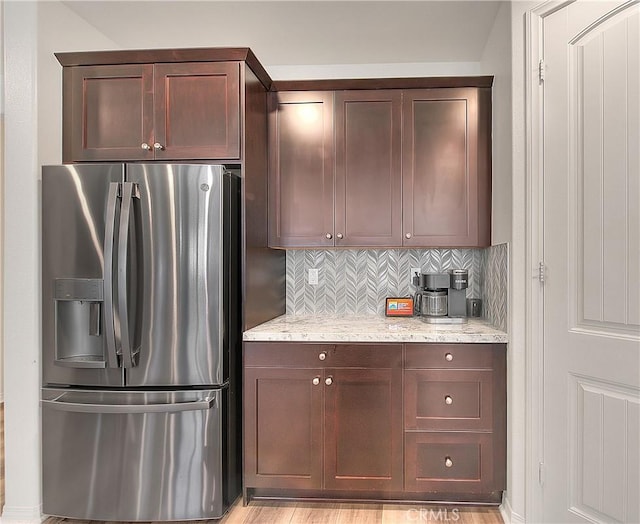 kitchen featuring light stone counters, backsplash, dark brown cabinets, and stainless steel refrigerator with ice dispenser