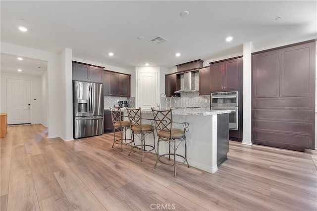 kitchen with light wood finished floors, stainless steel appliances, an island with sink, and wall chimney range hood