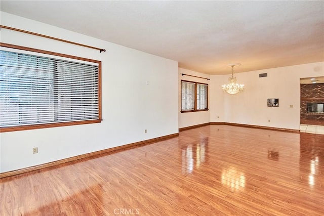 unfurnished living room with wood finished floors, visible vents, baseboards, a brick fireplace, and an inviting chandelier