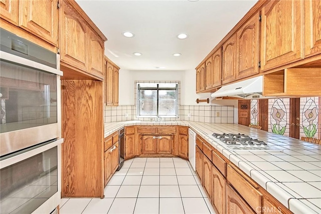 kitchen with white appliances, tasteful backsplash, tile counters, brown cabinets, and under cabinet range hood