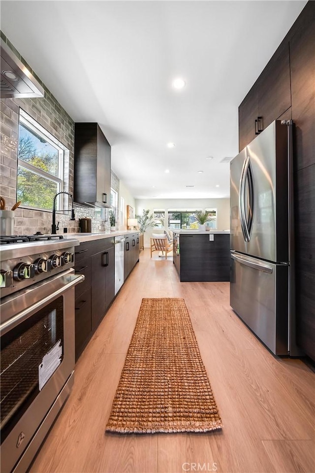 kitchen featuring tasteful backsplash, light countertops, appliances with stainless steel finishes, wall chimney range hood, and light wood-type flooring