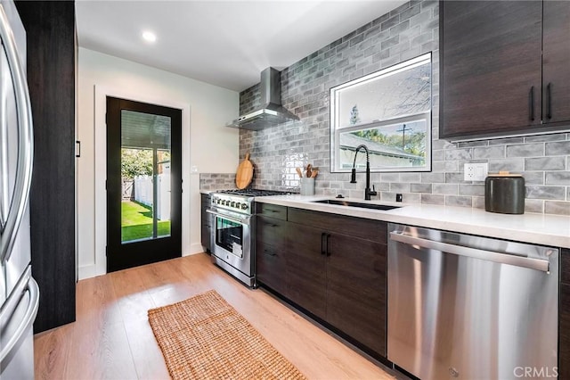 kitchen featuring wall chimney exhaust hood, stainless steel appliances, dark brown cabinets, light countertops, and a sink