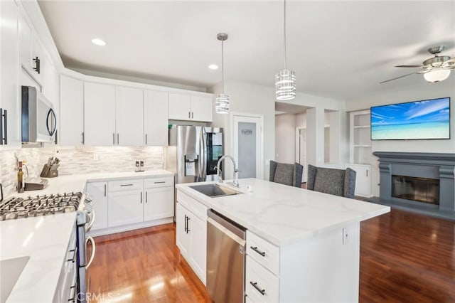 kitchen featuring wood finished floors, a center island with sink, a sink, stainless steel appliances, and a glass covered fireplace