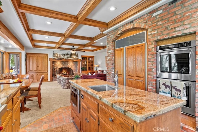 kitchen with double oven, a kitchen island with sink, a sink, a lit fireplace, and beamed ceiling