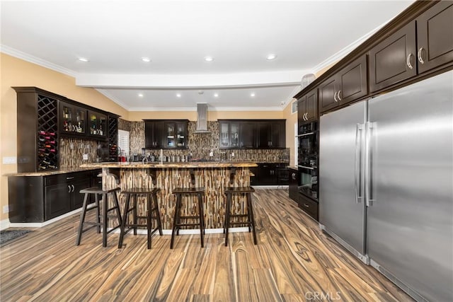kitchen featuring glass insert cabinets, a breakfast bar area, built in refrigerator, wood finished floors, and island range hood