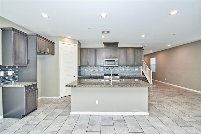 kitchen featuring stone countertops, a center island with sink, baseboards, stainless steel microwave, and dark brown cabinets