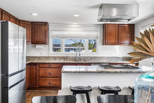 kitchen featuring light stone counters, a breakfast bar, freestanding refrigerator, a sink, and wall chimney range hood