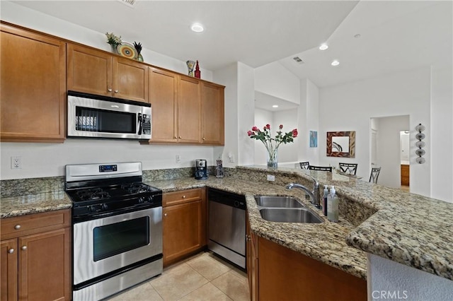 kitchen with brown cabinetry, stone counters, a peninsula, a sink, and appliances with stainless steel finishes