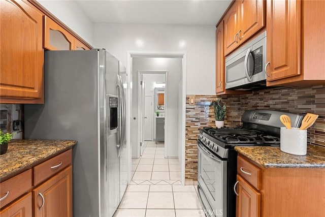 kitchen with brown cabinetry, decorative backsplash, dark stone countertops, appliances with stainless steel finishes, and light tile patterned flooring