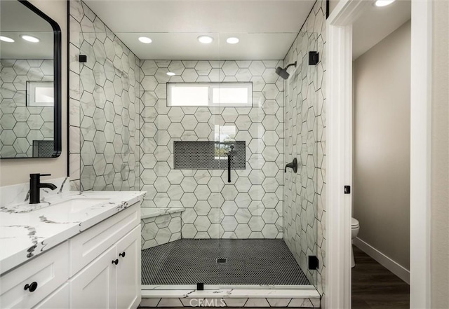 bathroom with toilet, vanity, wood finished floors, tiled shower, and baseboards