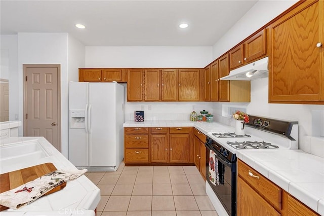 kitchen with tile counters, brown cabinetry, range with gas cooktop, under cabinet range hood, and white fridge with ice dispenser