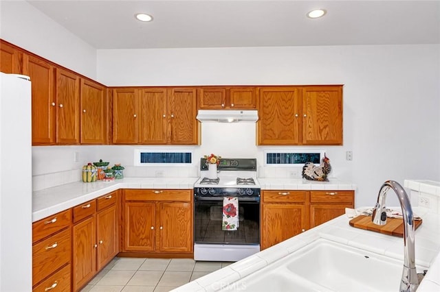 kitchen featuring tile countertops, gas stove, freestanding refrigerator, a sink, and under cabinet range hood