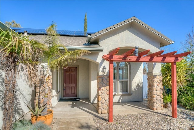 view of exterior entry with a shingled roof, a pergola, roof mounted solar panels, and stucco siding