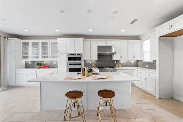 kitchen with visible vents, an island with sink, light countertops, under cabinet range hood, and double oven