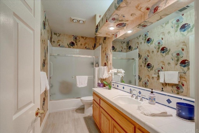 bathroom with bath / shower combo with glass door, visible vents, toilet, vanity, and wallpapered walls