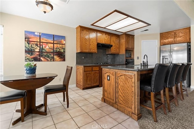 kitchen with dark countertops, decorative backsplash, brown cabinetry, under cabinet range hood, and stainless steel fridge with ice dispenser