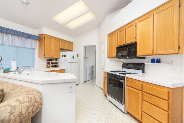 kitchen with tile countertops, freestanding refrigerator, black microwave, washer and dryer, and gas range