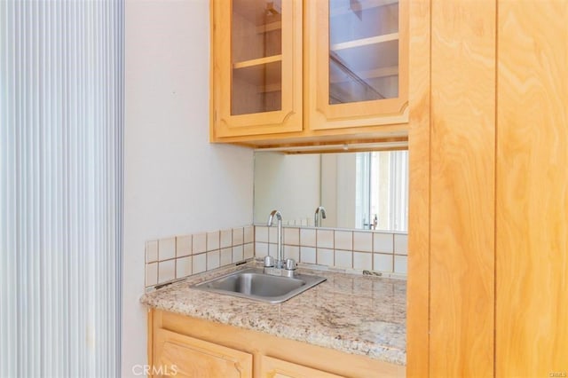 kitchen with light stone counters, backsplash, a sink, light brown cabinetry, and glass insert cabinets