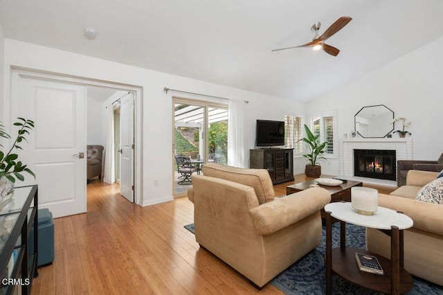 living room featuring a ceiling fan, a fireplace, light wood-style floors, baseboards, and vaulted ceiling