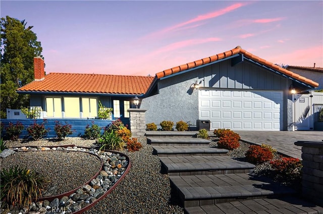 rear view of house featuring decorative driveway, a tile roof, a chimney, stucco siding, and an attached garage