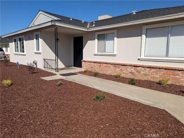 entrance to property featuring brick siding, roof with shingles, and stucco siding