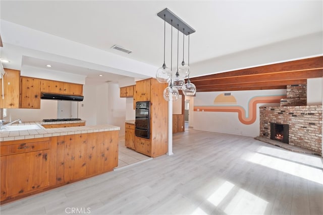 kitchen featuring visible vents, tile counters, light wood-style flooring, a peninsula, and a brick fireplace