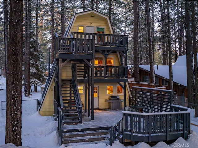 snow covered rear of property with stairway, a gambrel roof, a wooden deck, and fence