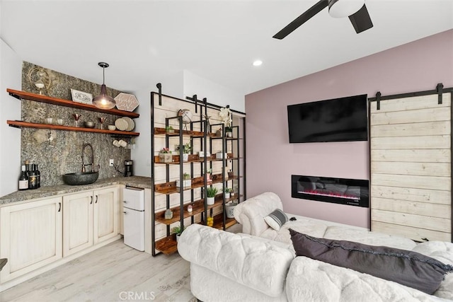 living area featuring ceiling fan, a barn door, indoor wet bar, light wood-style flooring, and a glass covered fireplace