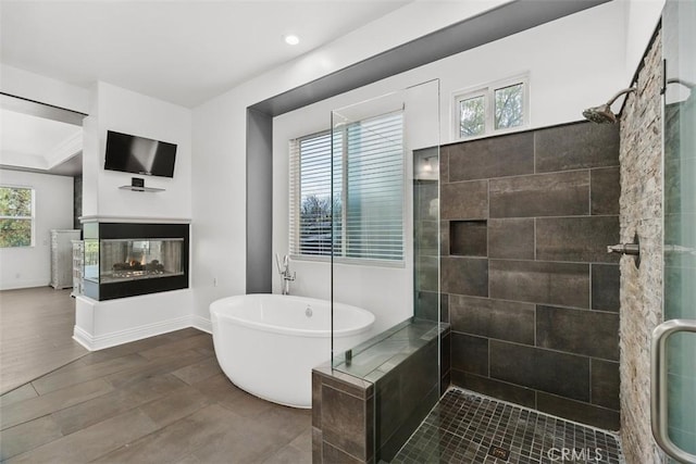 full bath with baseboards, a multi sided fireplace, tiled shower, wood finished floors, and a soaking tub