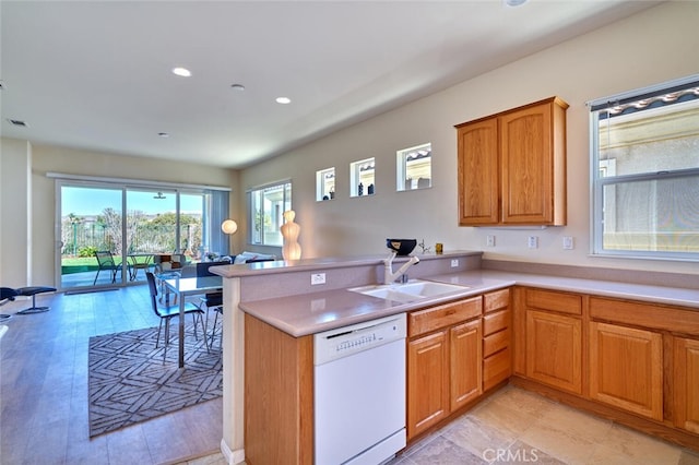 kitchen featuring white dishwasher, a peninsula, a sink, light countertops, and brown cabinets