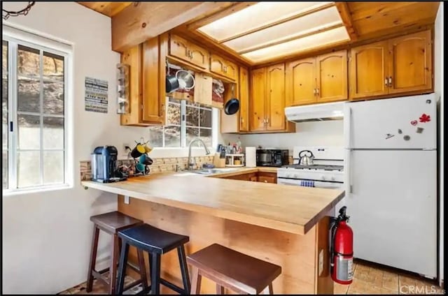 kitchen featuring brown cabinets, a sink, a peninsula, white appliances, and under cabinet range hood