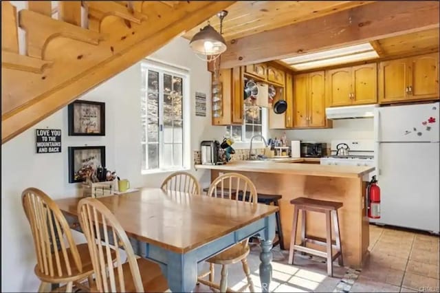 kitchen featuring freestanding refrigerator, a sink, a peninsula, beamed ceiling, and under cabinet range hood