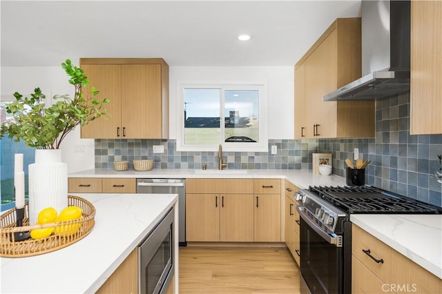 kitchen featuring light brown cabinets, a sink, light wood-style floors, appliances with stainless steel finishes, and wall chimney range hood