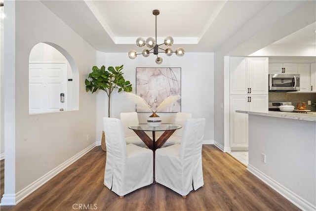 dining room with dark wood-style floors, a tray ceiling, arched walkways, a notable chandelier, and baseboards