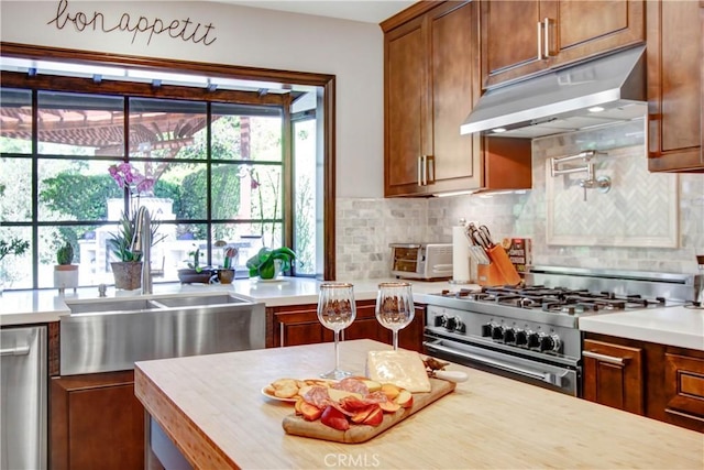 kitchen with under cabinet range hood, stainless steel appliances, light countertops, backsplash, and brown cabinets