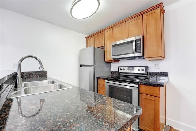 kitchen featuring stainless steel appliances, a sink, baseboards, brown cabinetry, and dark stone countertops