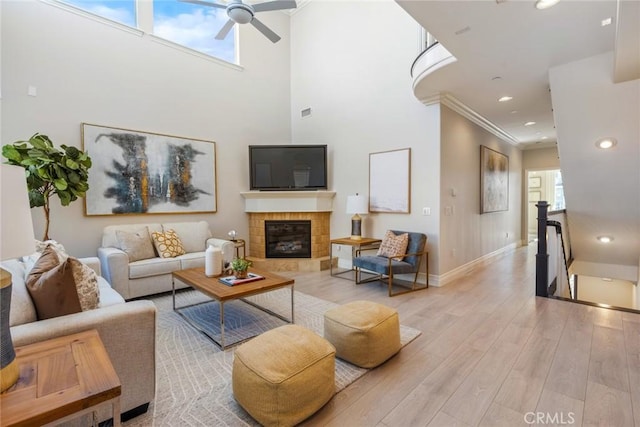 living area featuring crown molding, baseboards, light wood-style floors, a tiled fireplace, and a ceiling fan