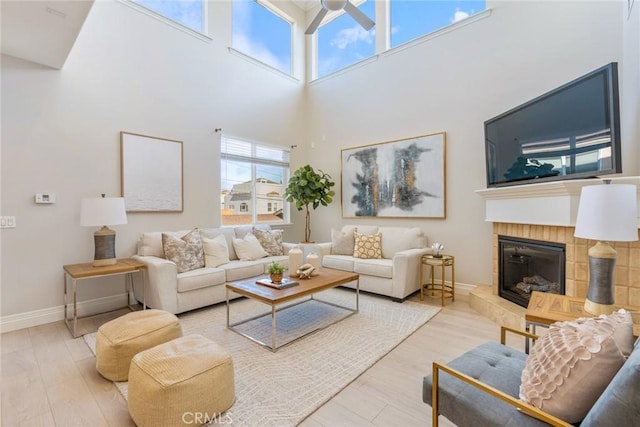 living room featuring baseboards, wood finished floors, a tiled fireplace, a ceiling fan, and a high ceiling